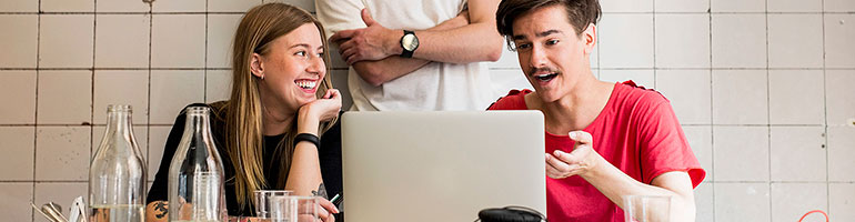 En tjej och en kille sitter vid en dator och jobbar. Bakom dem står en tredje person med armarna i kors.